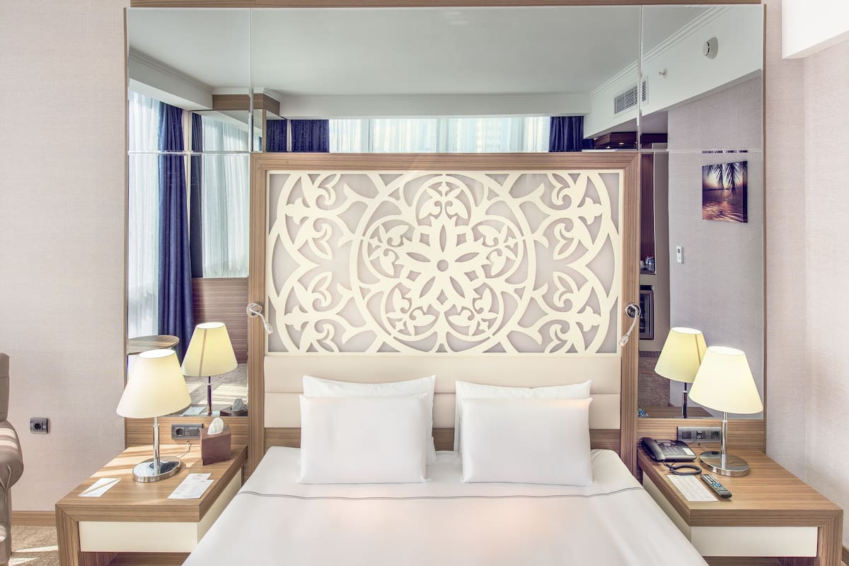 A hotel room with a large bed, two bedside tables with lamps, a decorative headboard, a mirror, and a view of the city through the window.
