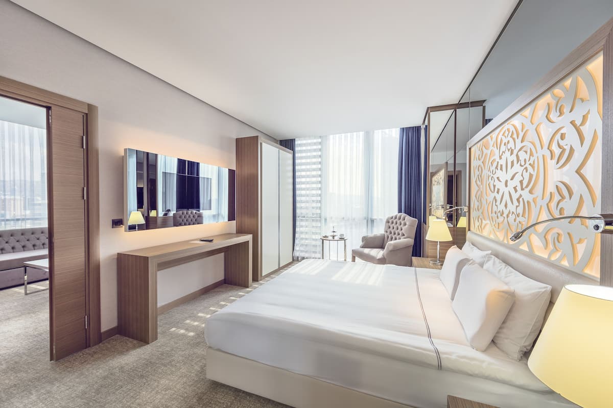 A modern hotel room with a large bed, a desk, a chair, a small table, and a decorative wall panel.