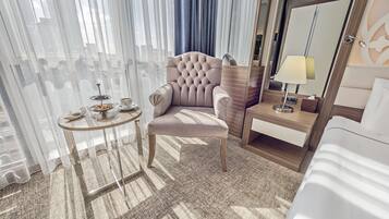 Una camera d'albergo con una poltrona trapuntata, un comodino con una lampada e vista attraverso tende leggere.