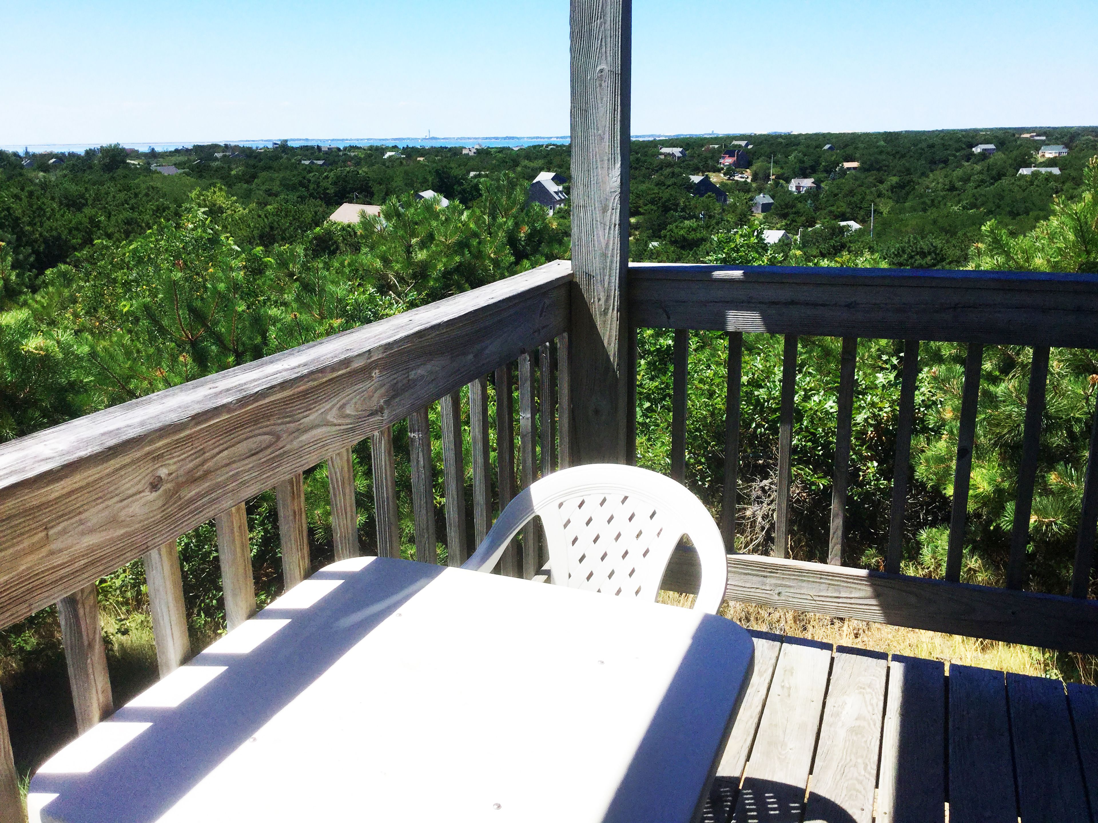 A wooden deck with a white plastic chair, overlooking a forested area with houses in the distance.