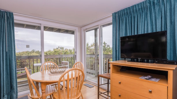 A room with a dining table, chairs, a television on a wooden stand, and large windows with curtains. - Cape View Motel (North Truro)
