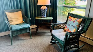 Two wicker chairs with cushions, a small table with a lamp, and a view of greenery outside. - Cape View Motel (North Truro)