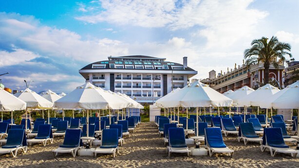 A beach with rows of blue lounge chairs and white umbrellas, a hotel building in the background, and a palm tree. - White Gold Hotel & Spa (Alanya)