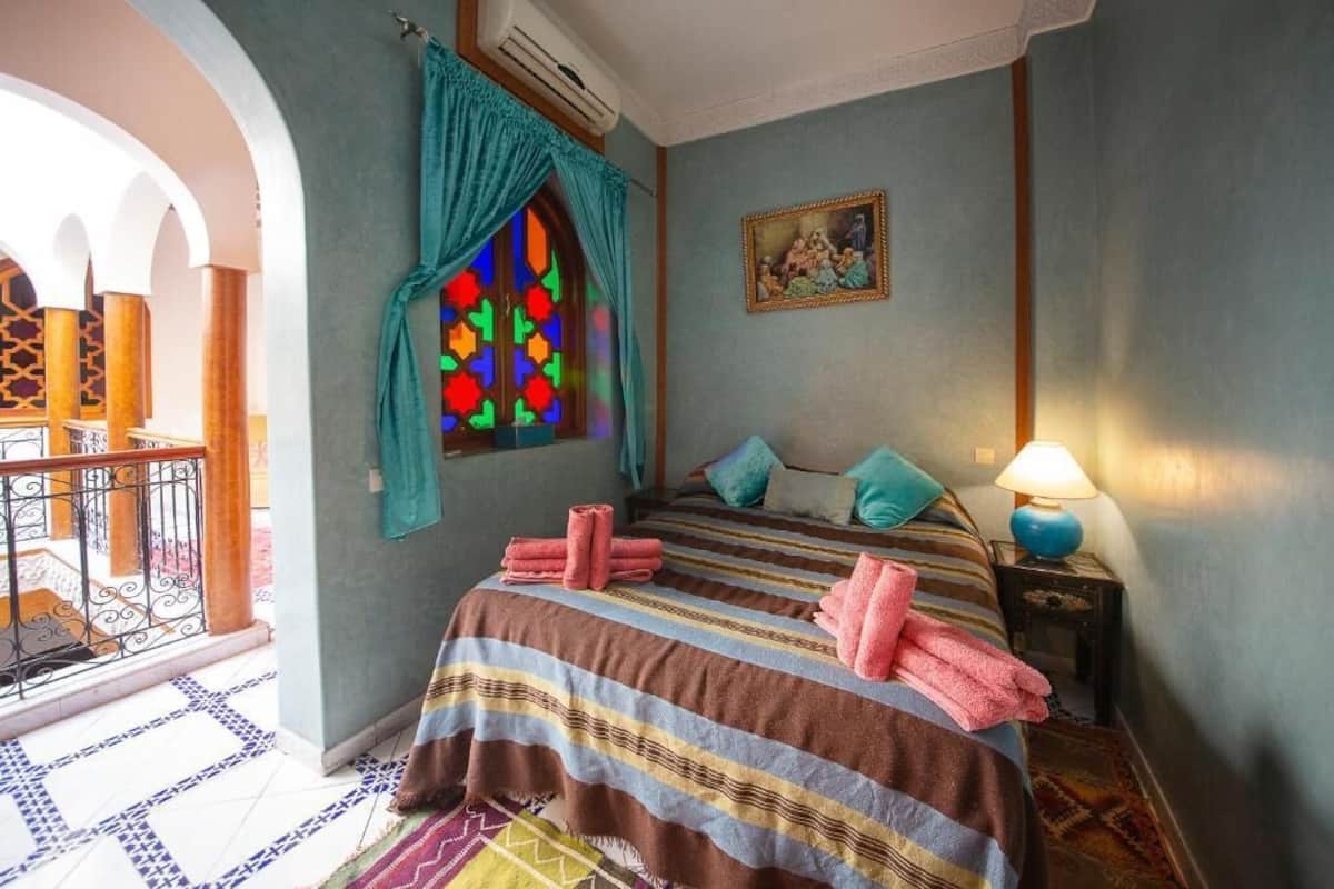 A bedroom with a bed, a window with colorful stained glass, a painting, and a balcony with a railing.