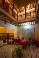 Uma área de refeições com mesas redondas cobertas por toalhas vermelhas, com piso estampado e alto teto com vigas de madeira.