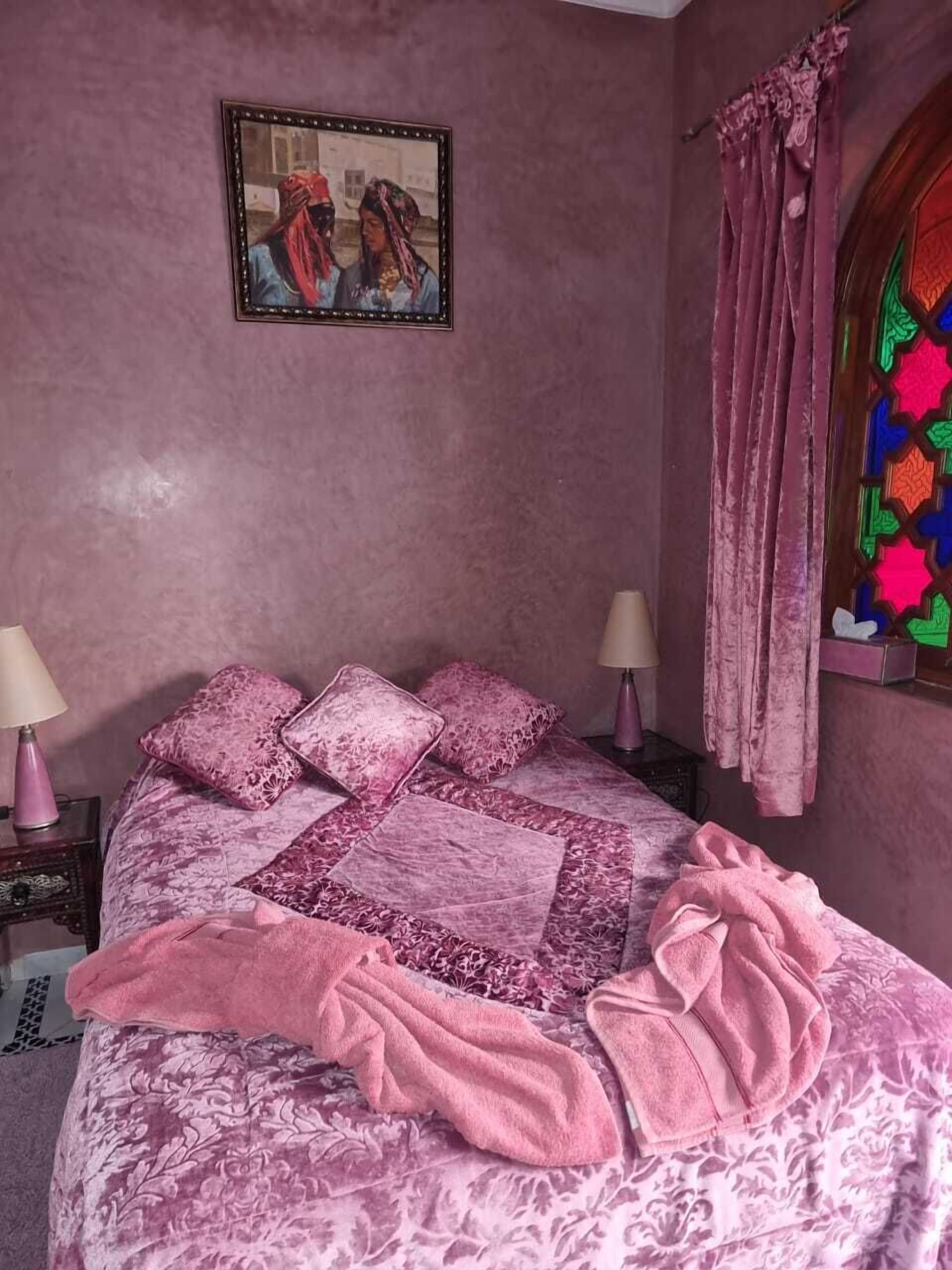 Ein Schlafzimmer mit einem Bett, rosa Bettwäsche, einem Nachttisch, einer Lampe und einem Fenster mit Vorhängen.