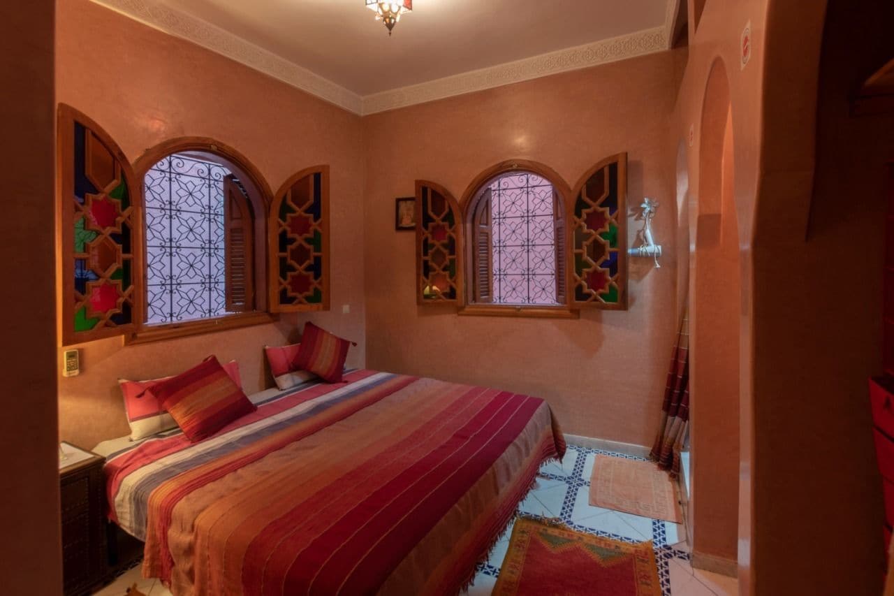 A bedroom with a large bed, two windows with decorative grills, and a chandelier.