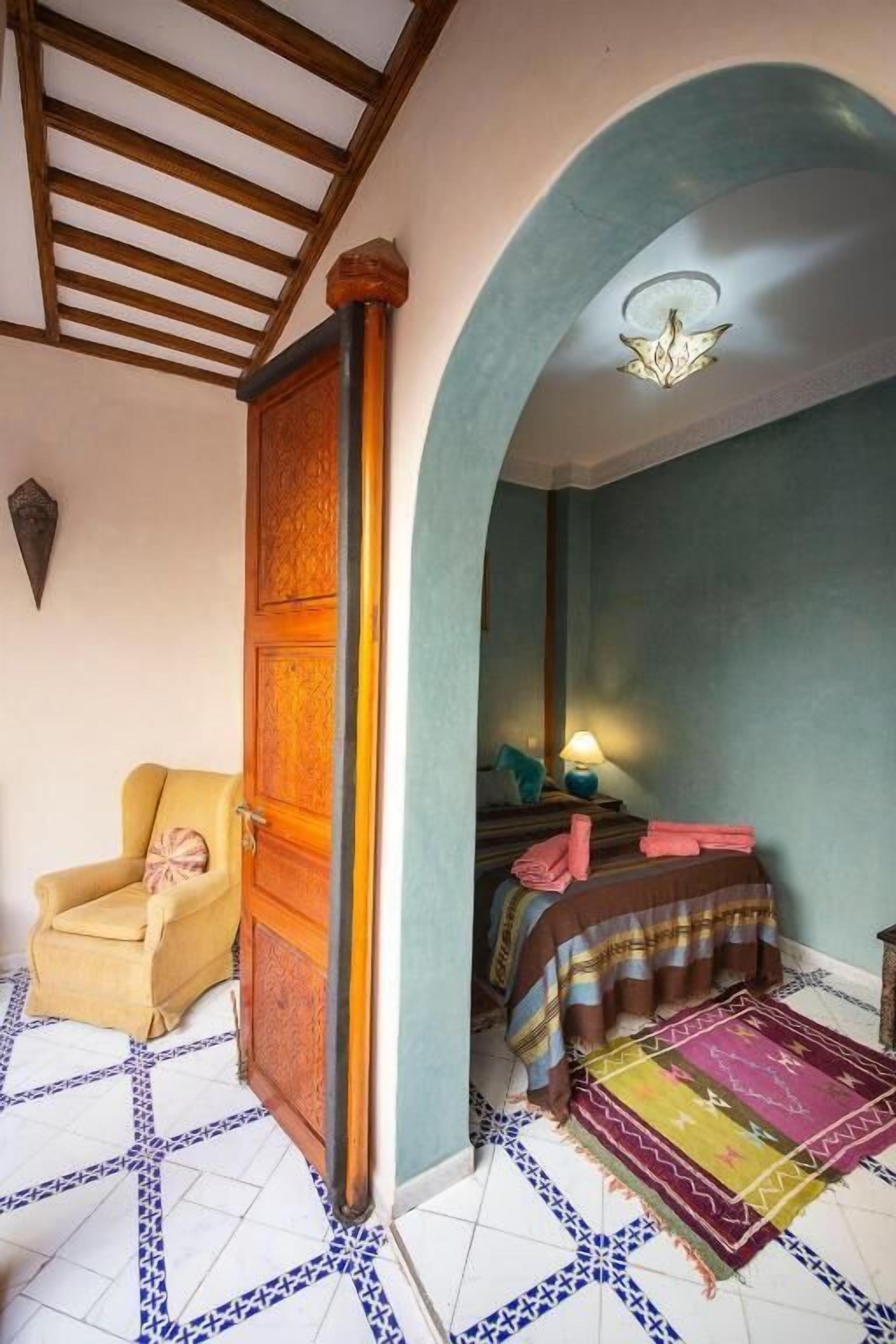 A room with a wooden door, a chair, a bed with a striped blanket, and a patterned rug.