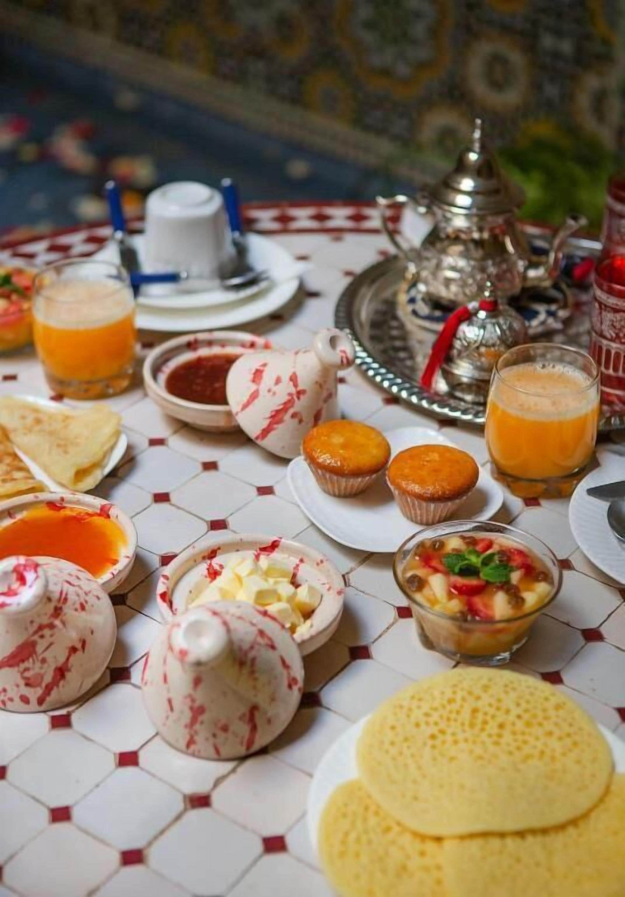 Une table recouverte de carreaux, dressée pour le petit-déjeuner, avec différents plats, notamment des crêpes, des muffins et du jus d’orange.