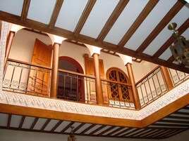 Uma varanda com guarda-corpo de madeira e detalhes em ferro decorativo, teto de madeira e vista do interior.