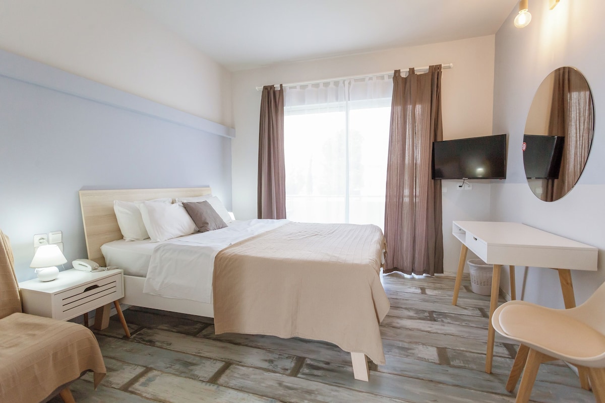 Una habitación de hotel con cama, mesita de noche, lámpara, silla, escritorio, televisor y cortinas.