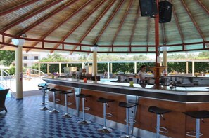 Uma área de bar ao ar livre coberta, com piso xadrez, uma piscina ao fundo e uma pessoa trabalhando no balcão.