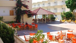 Un patio al aire libre con mesas y sillas redondas anaranjadas y blancas, un pabellón con techo rojo y un edificio con balcones al fondo.