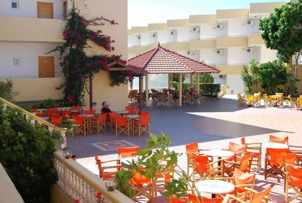 Un patio al aire libre con mesas y sillas redondas anaranjadas y blancas, un pabellón con techo rojo y un edificio con balcones al fondo.