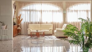 Sala de estar con un sofá beige, una mesa de centro redonda y un sillón blanco. Hay dos ventanas amplias con cortinas ligeras, un mueble de madera con adornos y una planta en maceta.