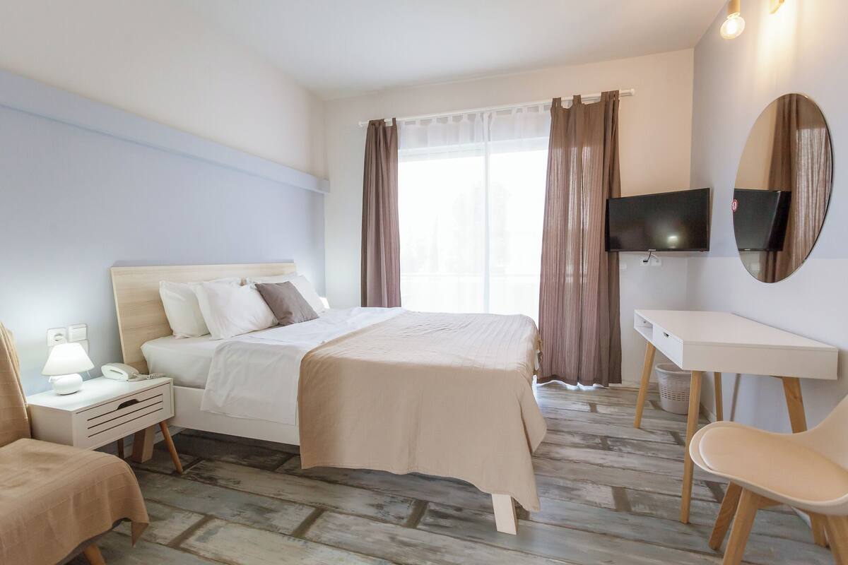 Una habitación de hotel con cama, mesita de noche, lámpara, silla, escritorio, televisor y cortinas.