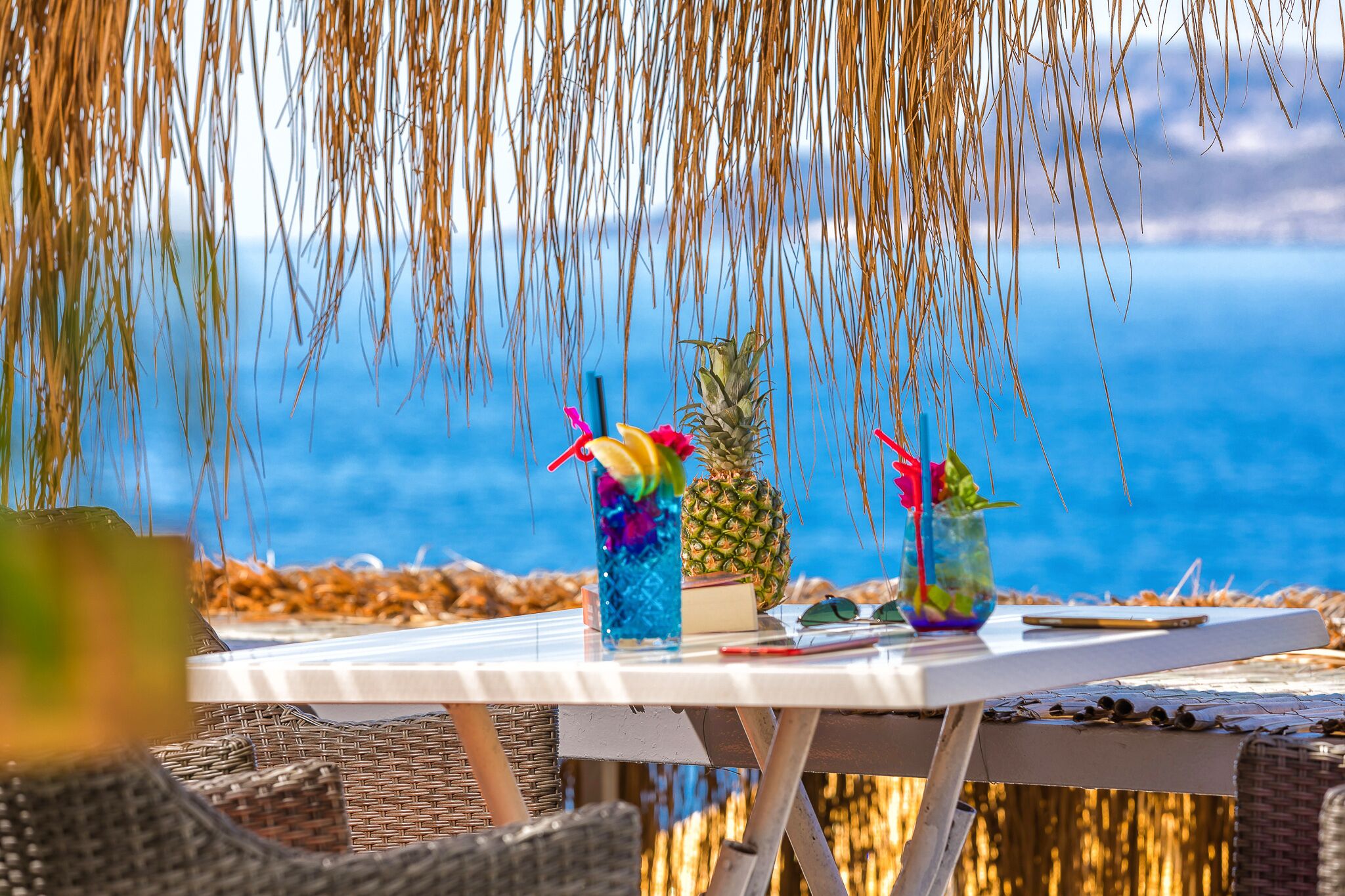 A table with drinks, a pineapple, and sunglasses, set against a scenic ocean view.