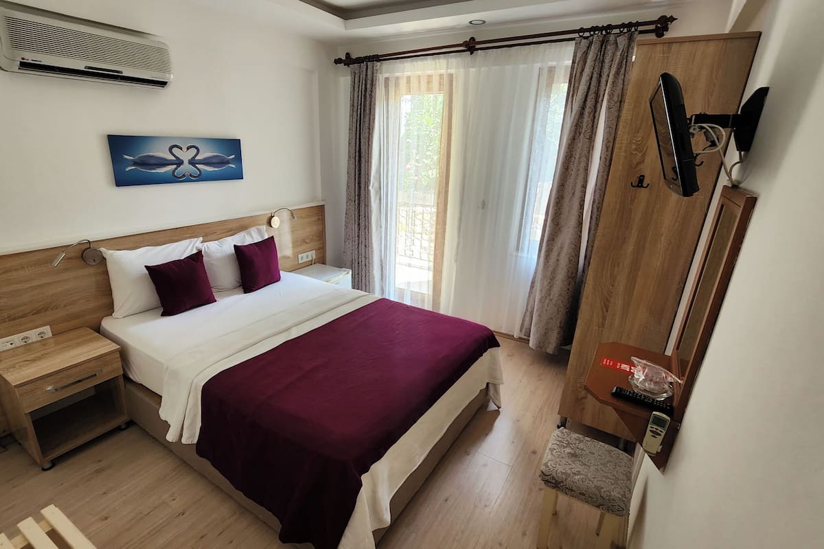 Un dormitorio con cabecera de madera, cama con ropa de cama blanca y burdeos, mesita de noche, armario de madera, televisor montado en la pared y ventana con cortinas.