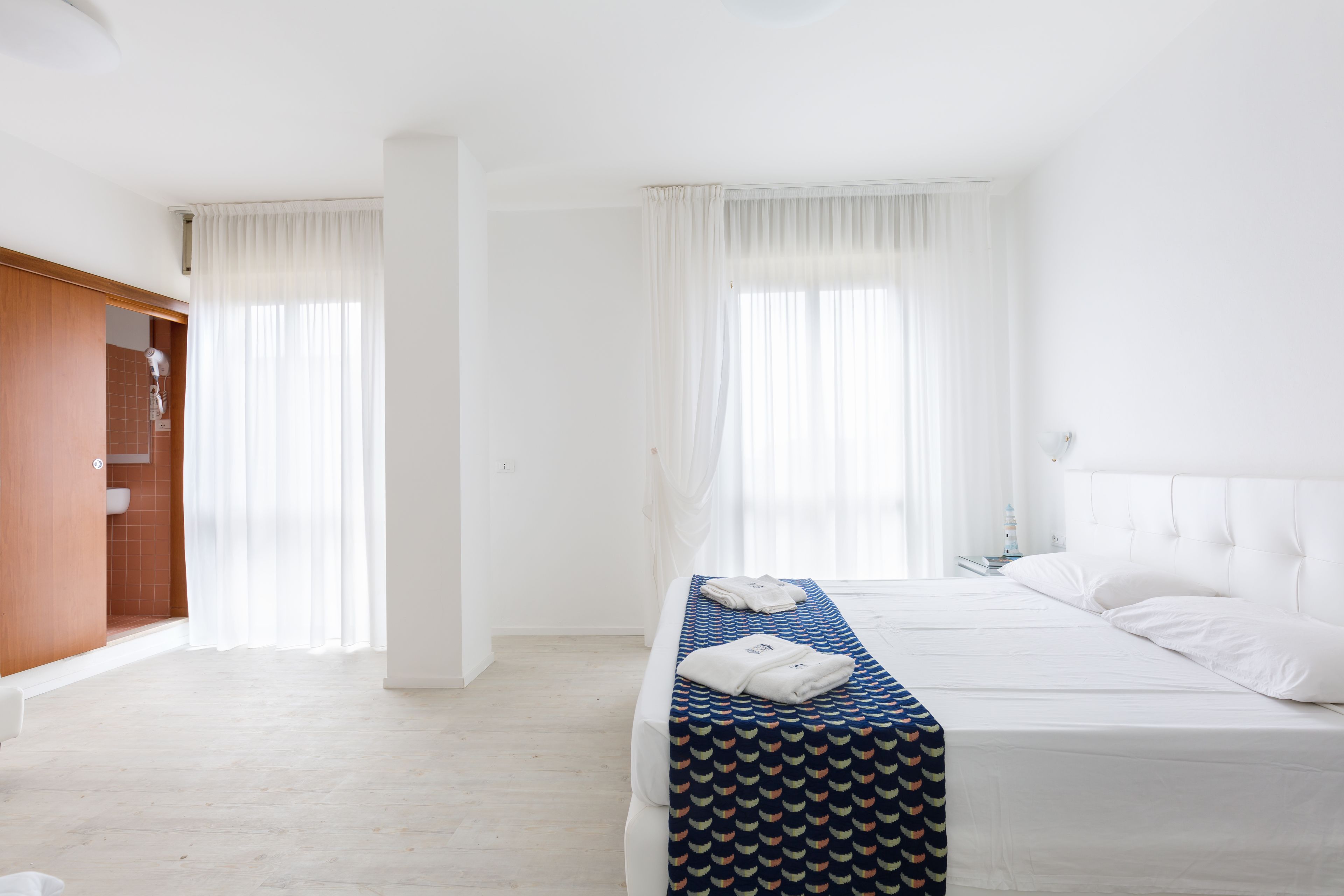 A hotel room with a bed, a bench with towels, a wooden wardrobe, and a window with curtains.