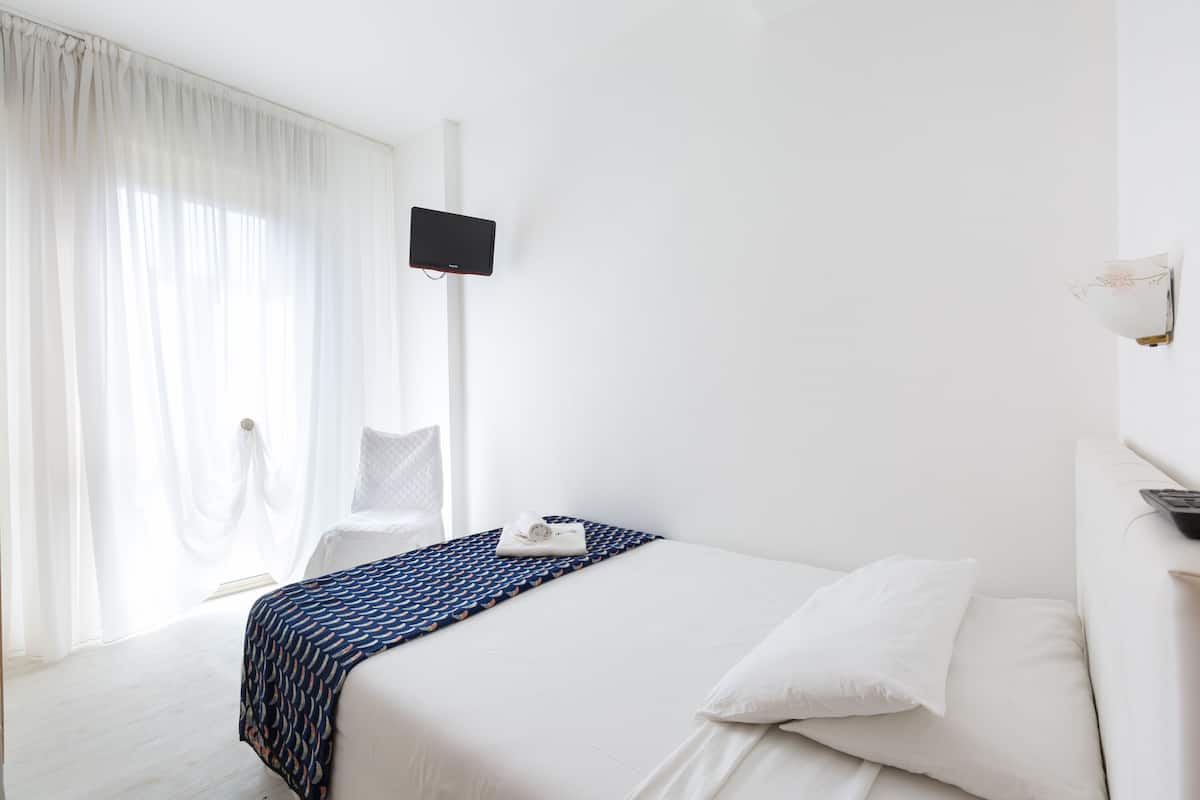 A hotel room with a bed, a TV mounted on the wall, a window with curtains, and a bedside table with a remote control.