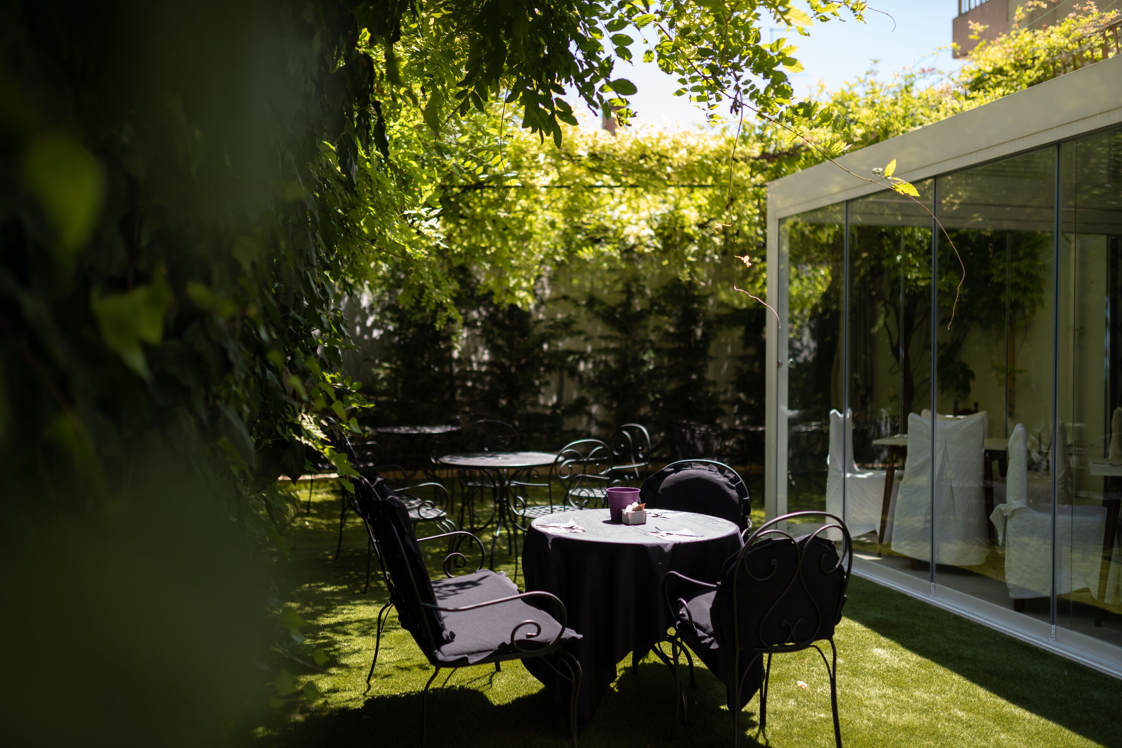 An outdoor patio with round tables and chairs, surrounded by lush greenery and a glass enclosure.