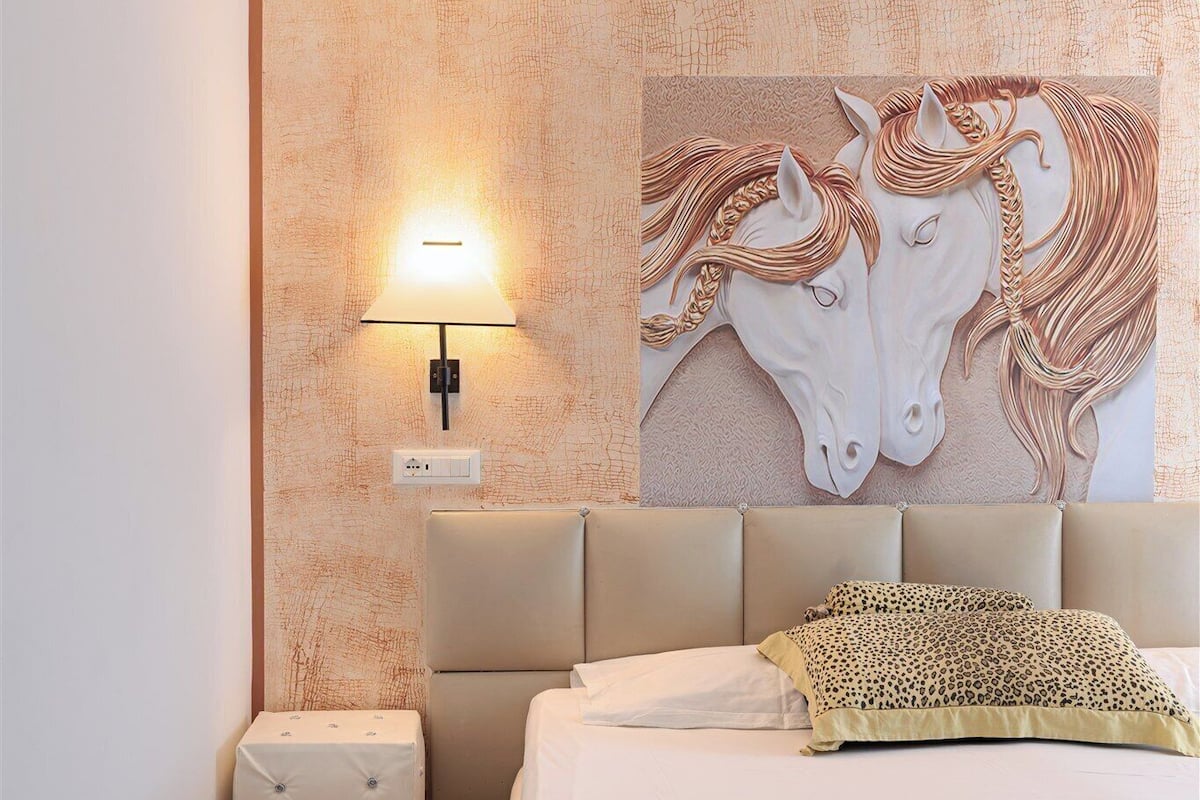 A bedroom with a bed, a nightstand, a wall-mounted lamp, and a large wall art of horses.