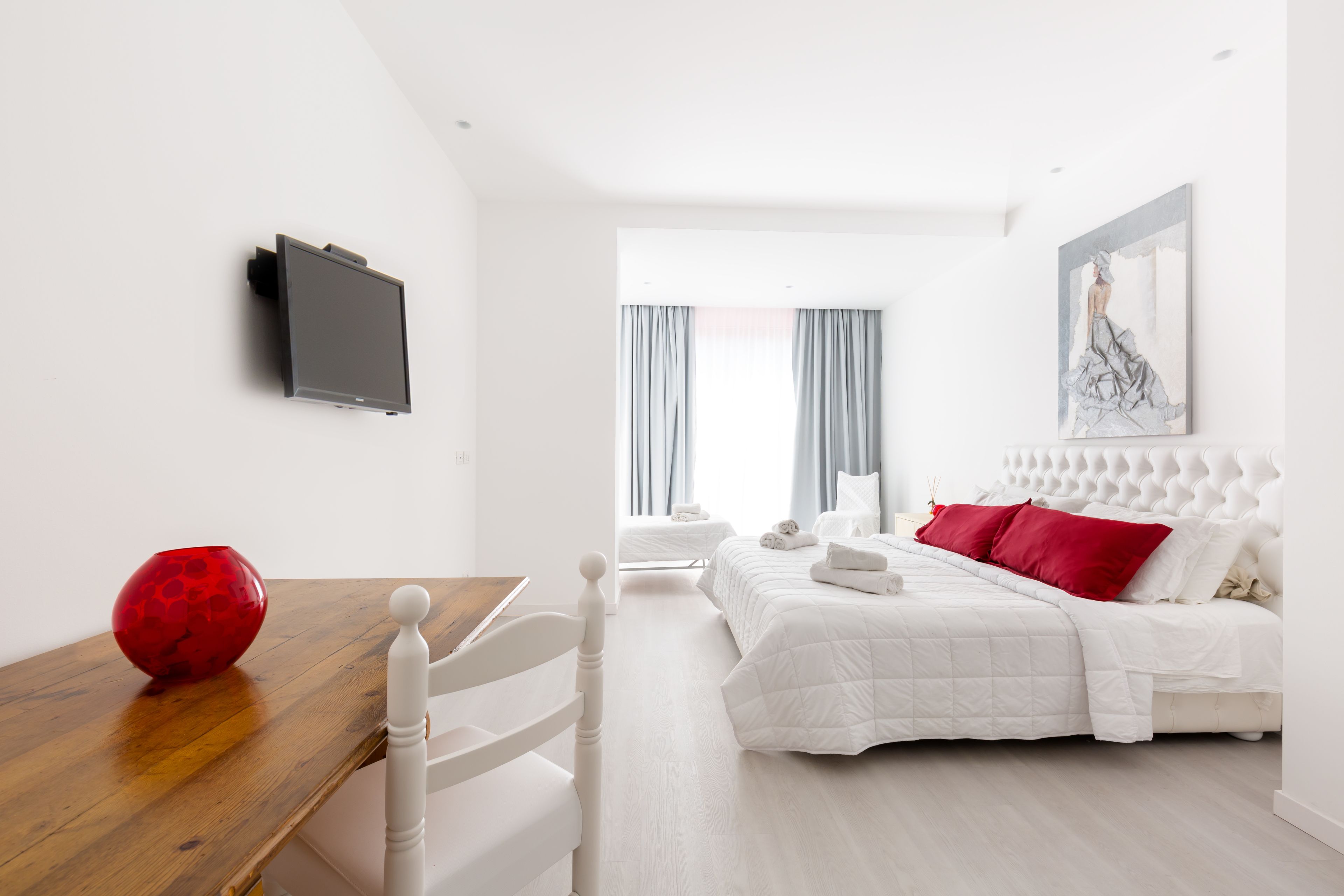 Moderni hotellihuone, jossa on puinen ruokapöytä, valkoinen sänky punaisilla tyynyillä, seinälle asennettu litteänäyttöinen televisio ja suuri ikkuna, jossa on verhot.