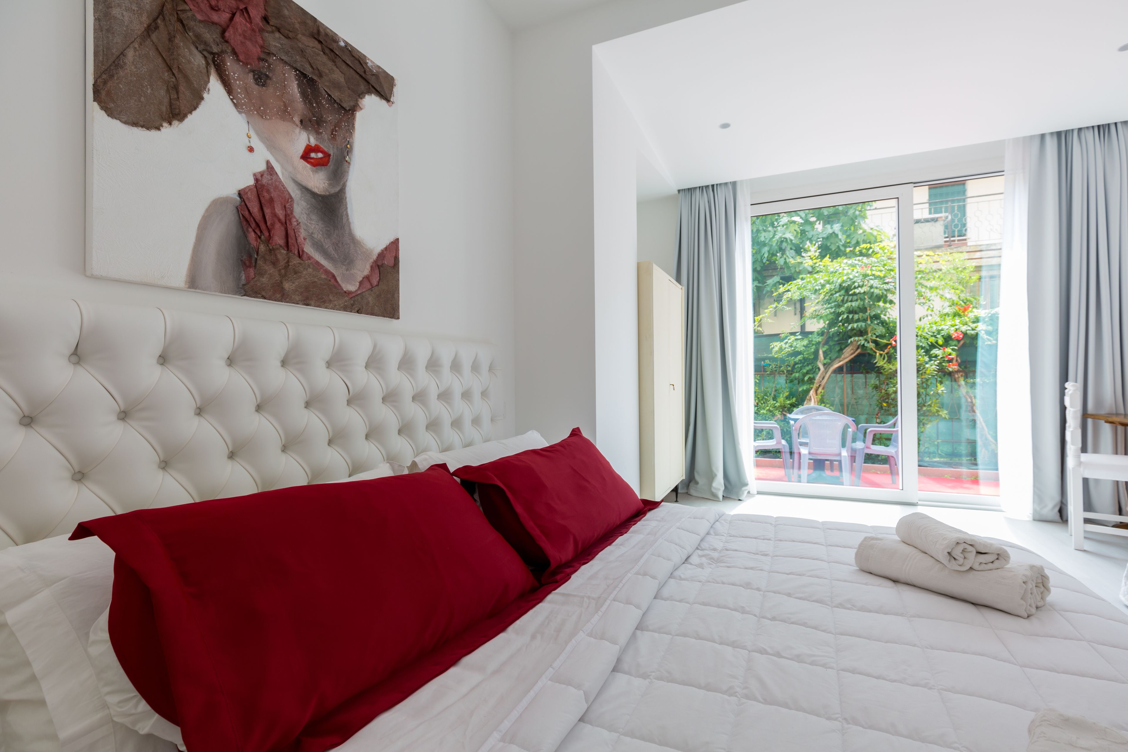 Ein Schlafzimmer mit einem großen Bett, einem roten und weißen Kopfkissen, einem weißen Kopfteil und einem Gemälde an der Wand.