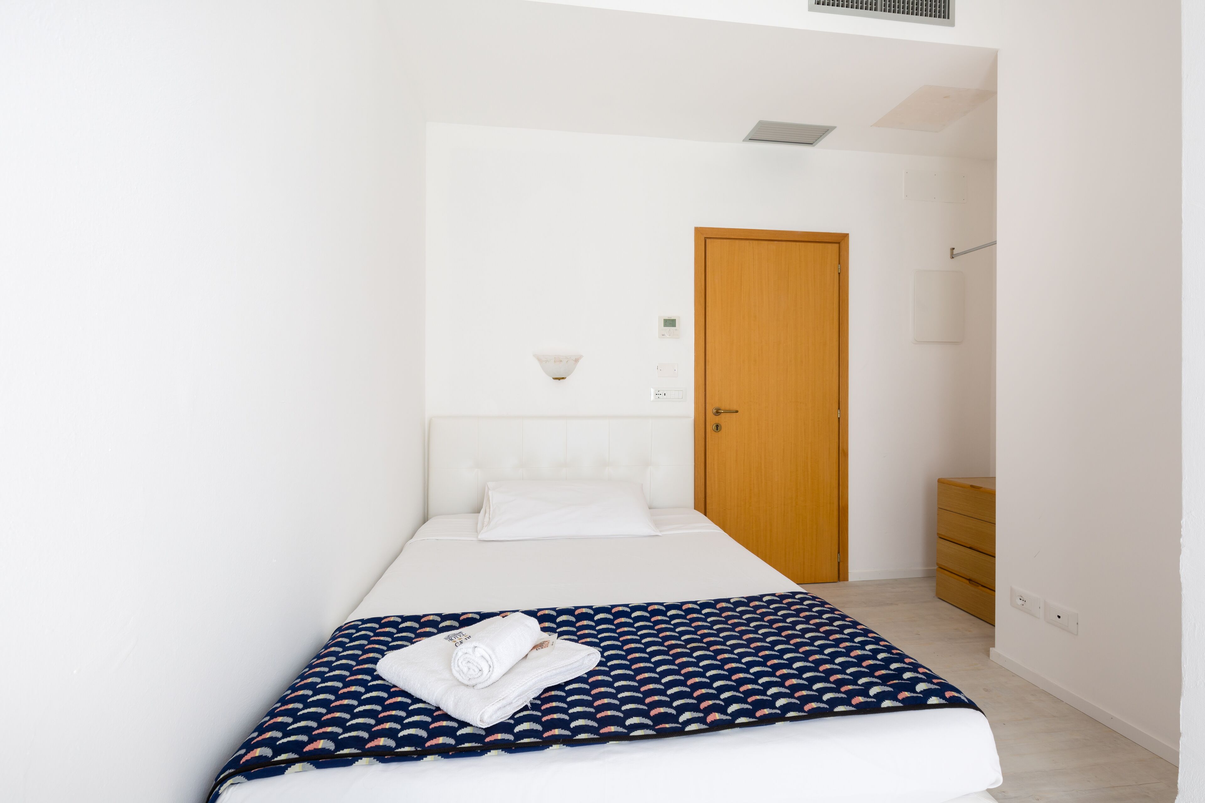 A neatly arranged bedroom with a bed, a wooden door, a wall-mounted light, and a small dresser.