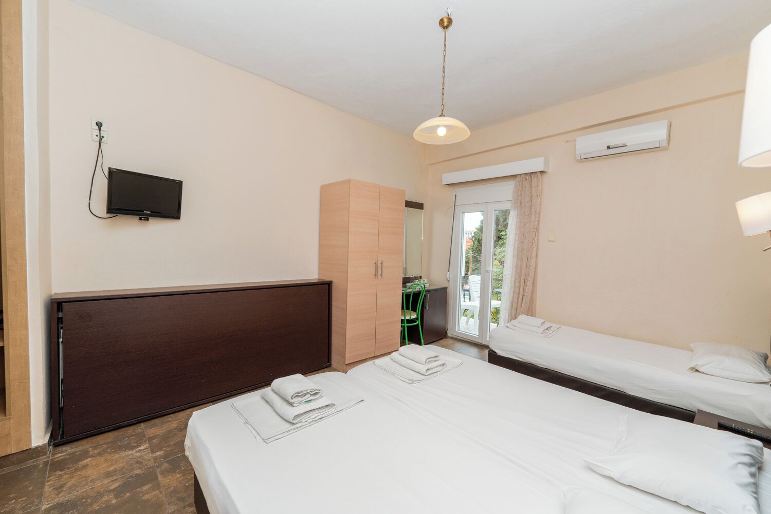 Habitación de hotel con dos camas, televisión, armario y balcón con una silla.