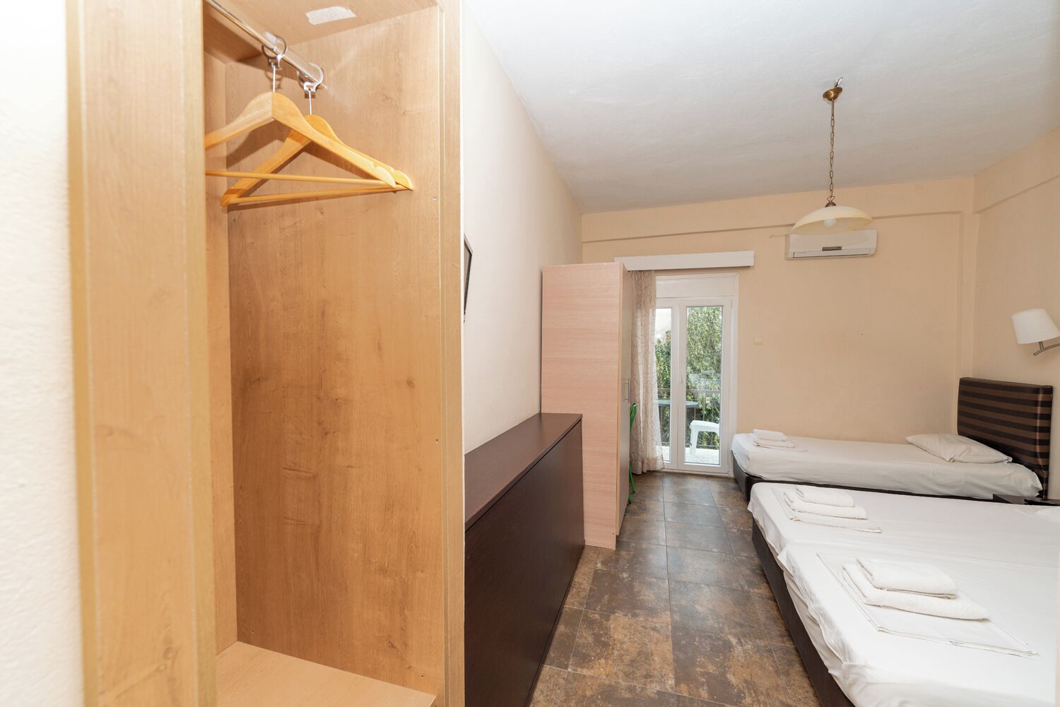 A hotel room with a wooden wardrobe, a desk, two beds, and a view of the outdoors through a glass door.