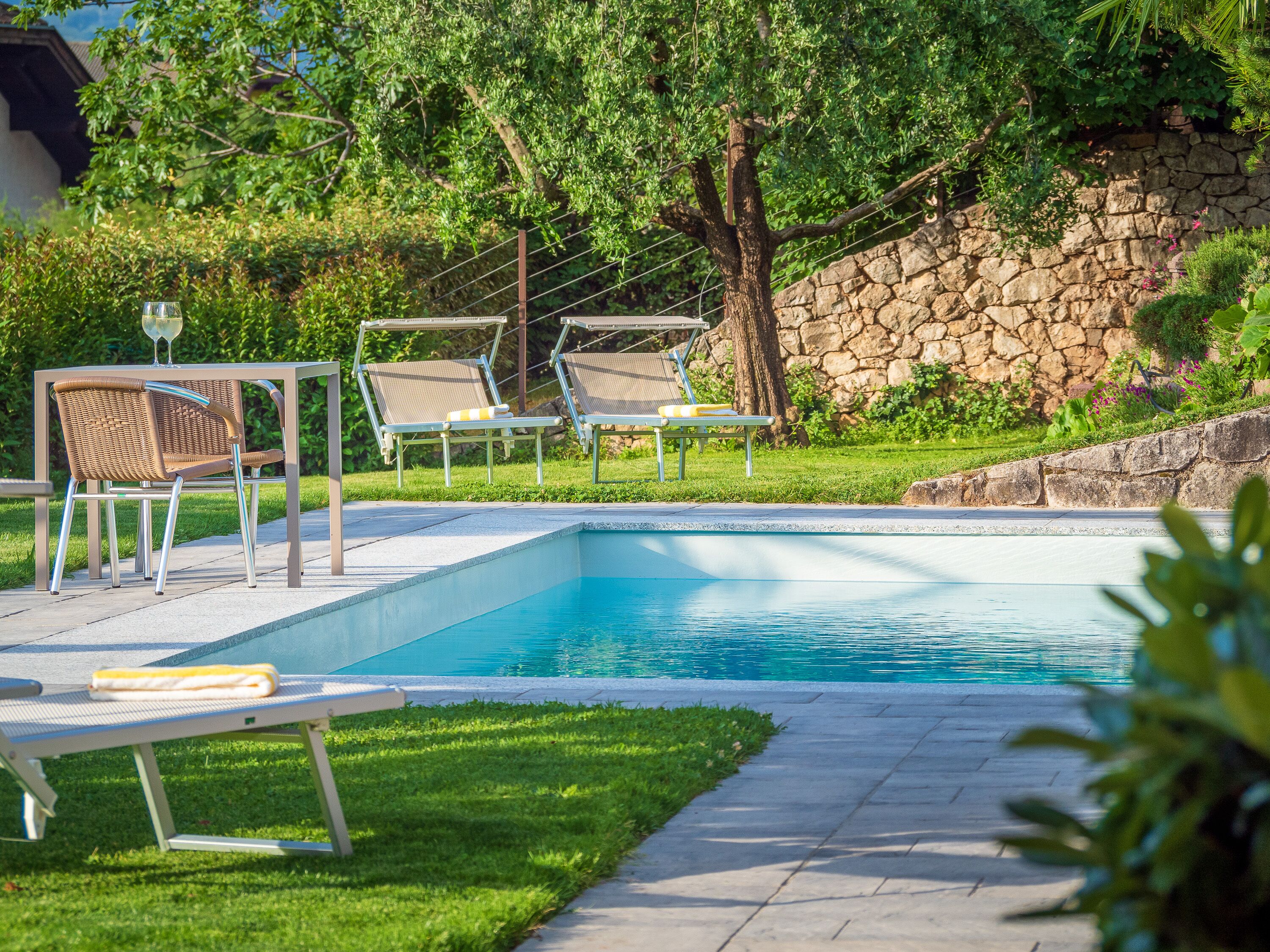 Piscina con sillas de descanso y una mesa, rodeada de vegetación y un muro de piedra.