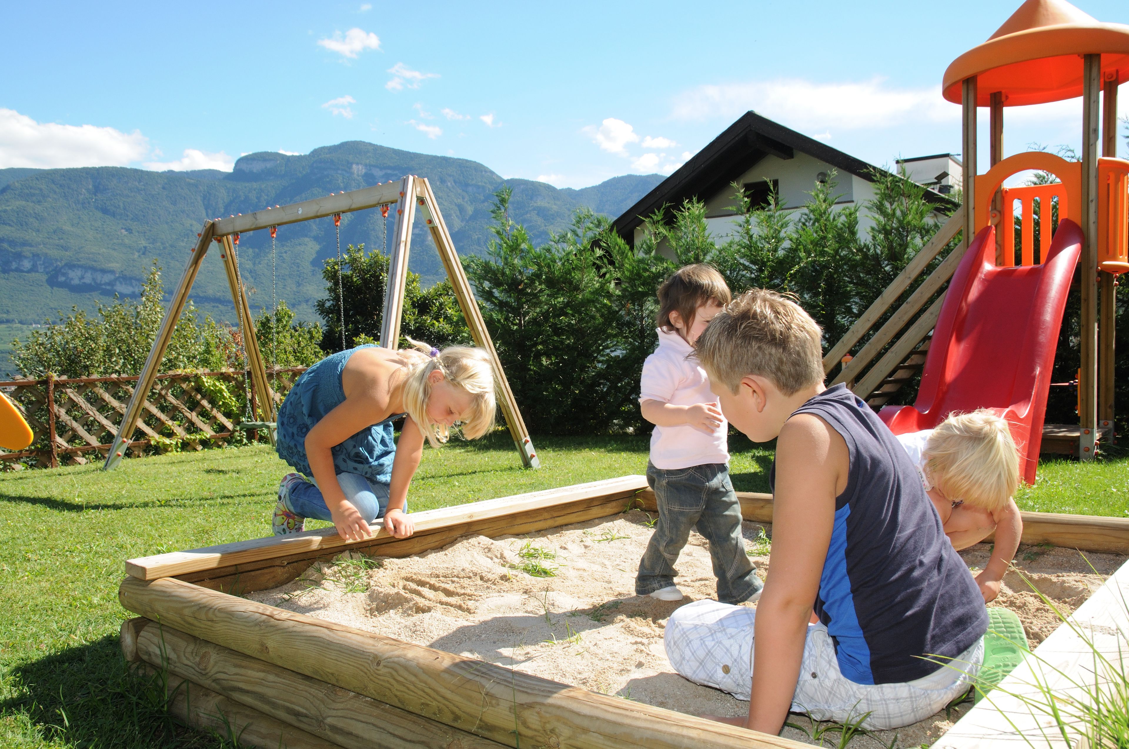 Children playing in a sandbox with a wooden swing set and a slide in the background.