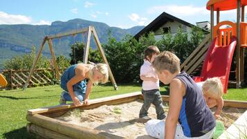 Niños jugando en un arenero con un columpio de madera y un tobogán al fondo.