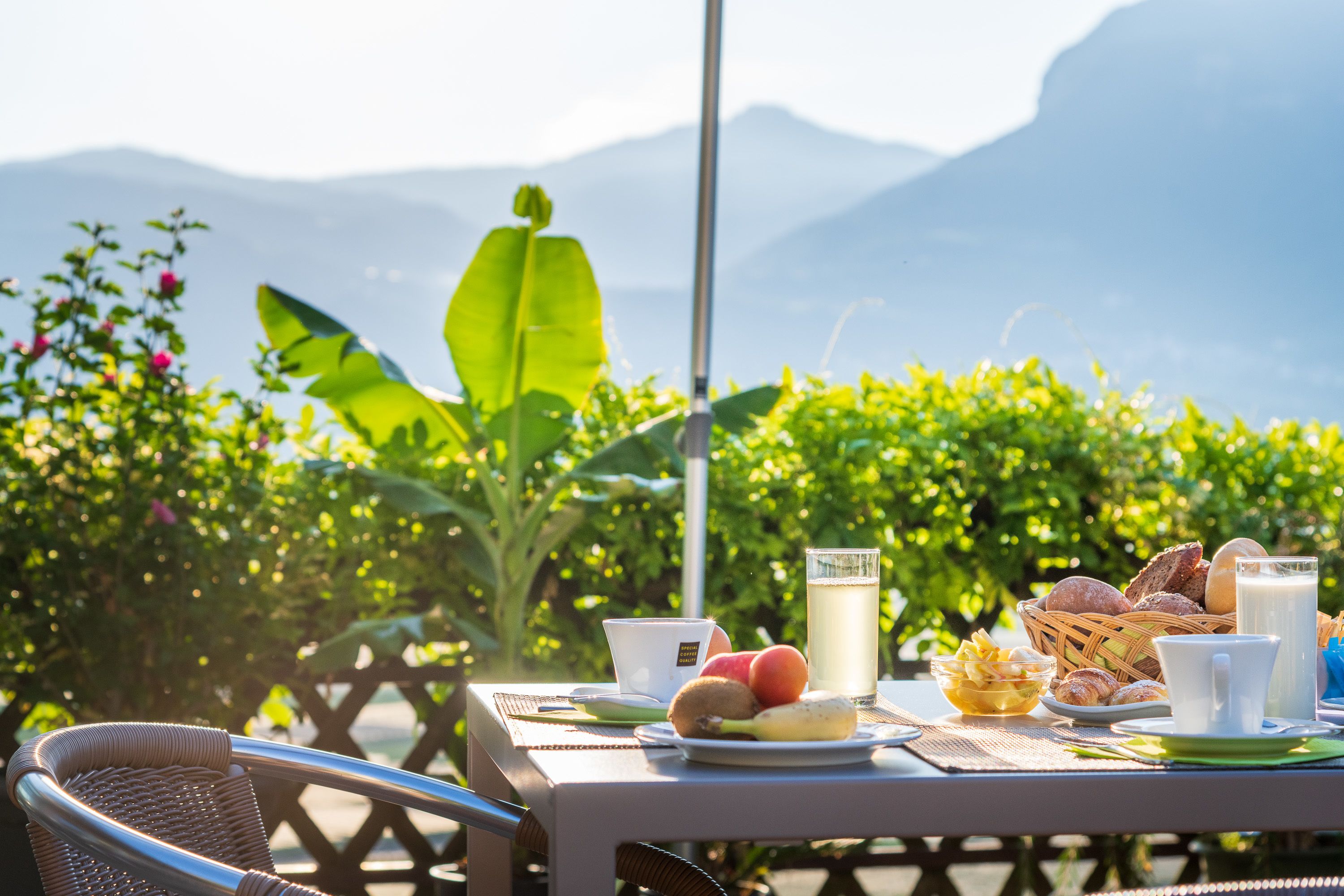 Mesa servida con alimentos para el desayuno, como frutas, pan y bebidas, con vistas a montañas al fondo.
