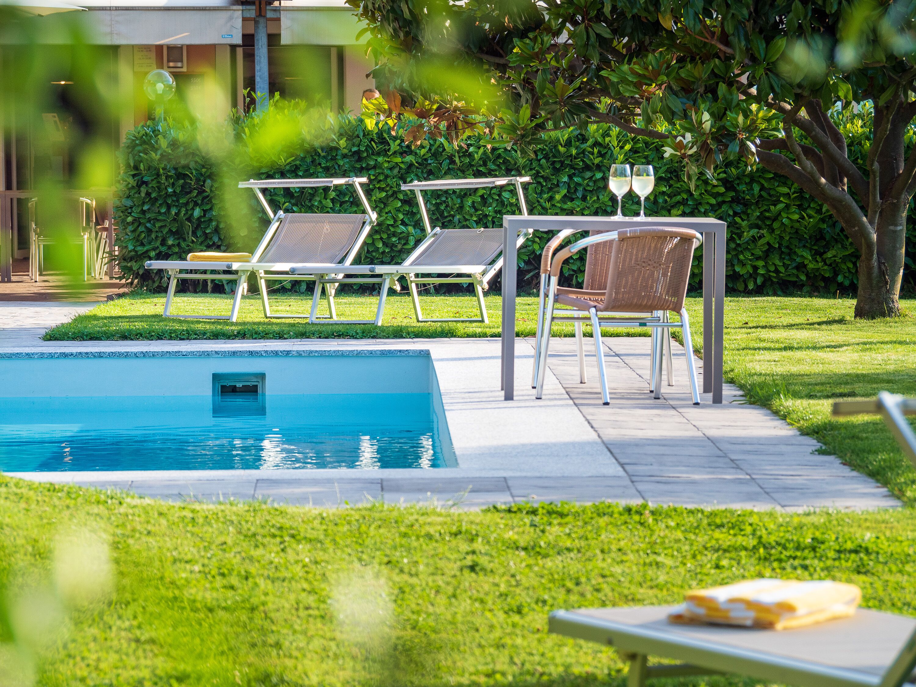 A poolside area with lounge chairs and a table, surrounded by greenery and a building in the background.