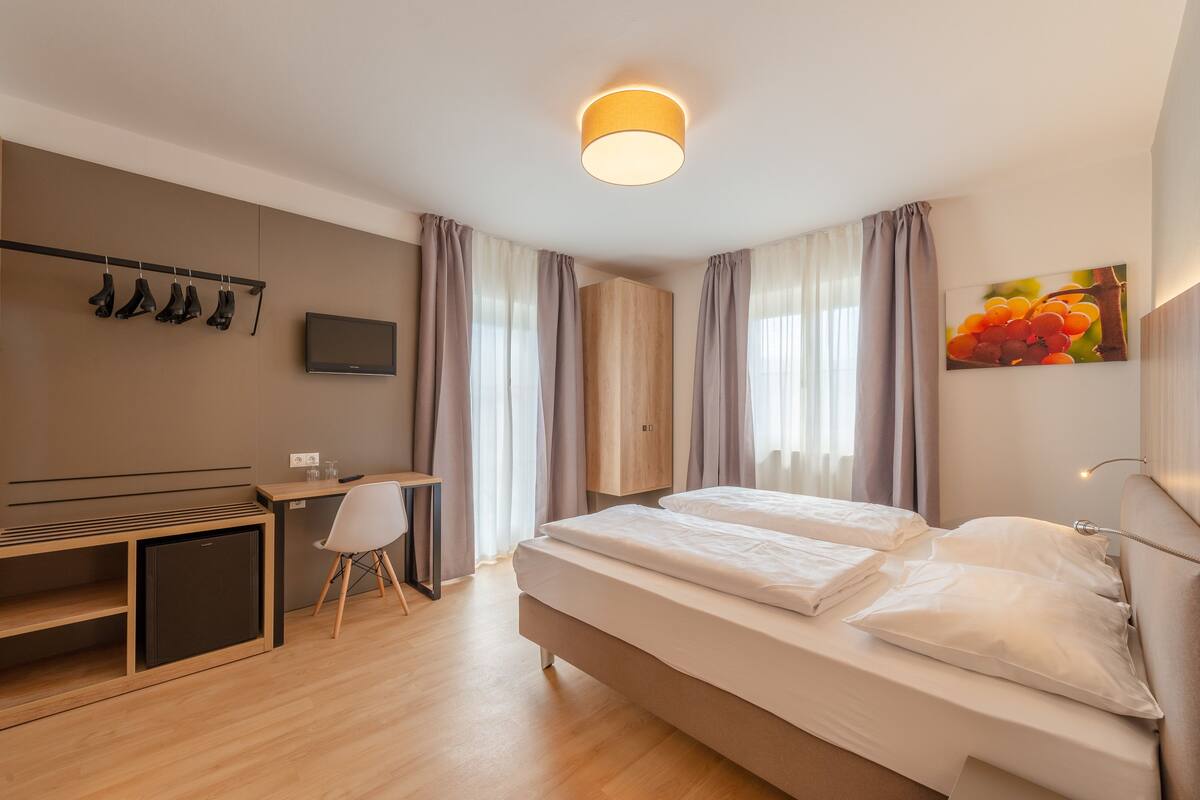 A modern hotel room with a bed, desk, chair, TV, and a coat rack with shoes.