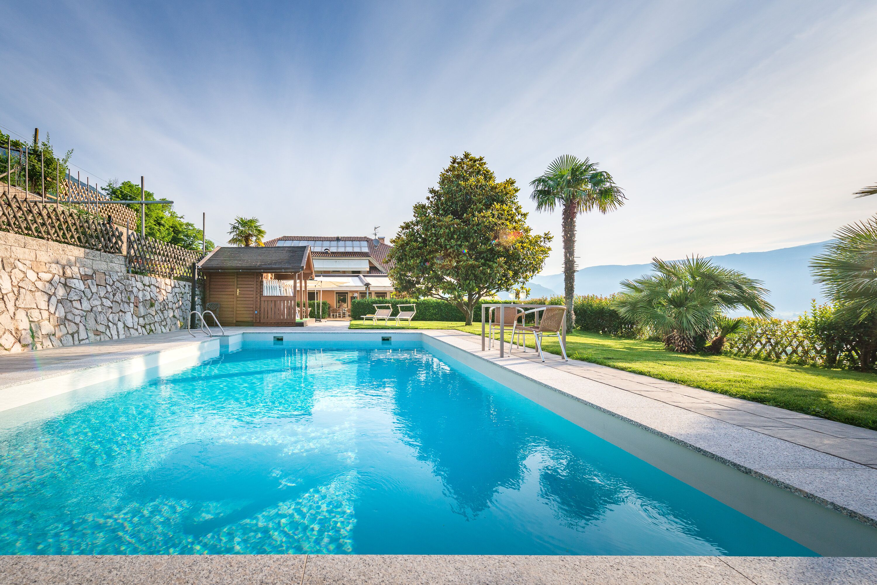 A swimming pool with a stone wall, a wooden deck, and a view of a house with a sloped roof.