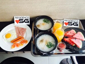 A breakfast tray with two bowls of porridge, a fried egg, ham, tomatoes, carrots, watermelon slices, and orange juice.