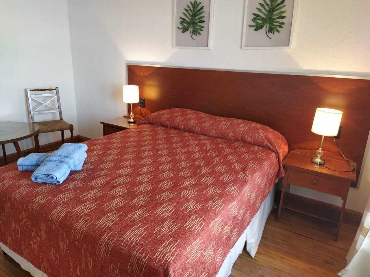 Un lit bien fait, recouvert d’une couvre-lit rouge à motifs, et deux serviettes pliées sont posées dessus. On peut voir deux tables de chevet avec des lampes, et une chaise est placée dans un coin de la pièce.