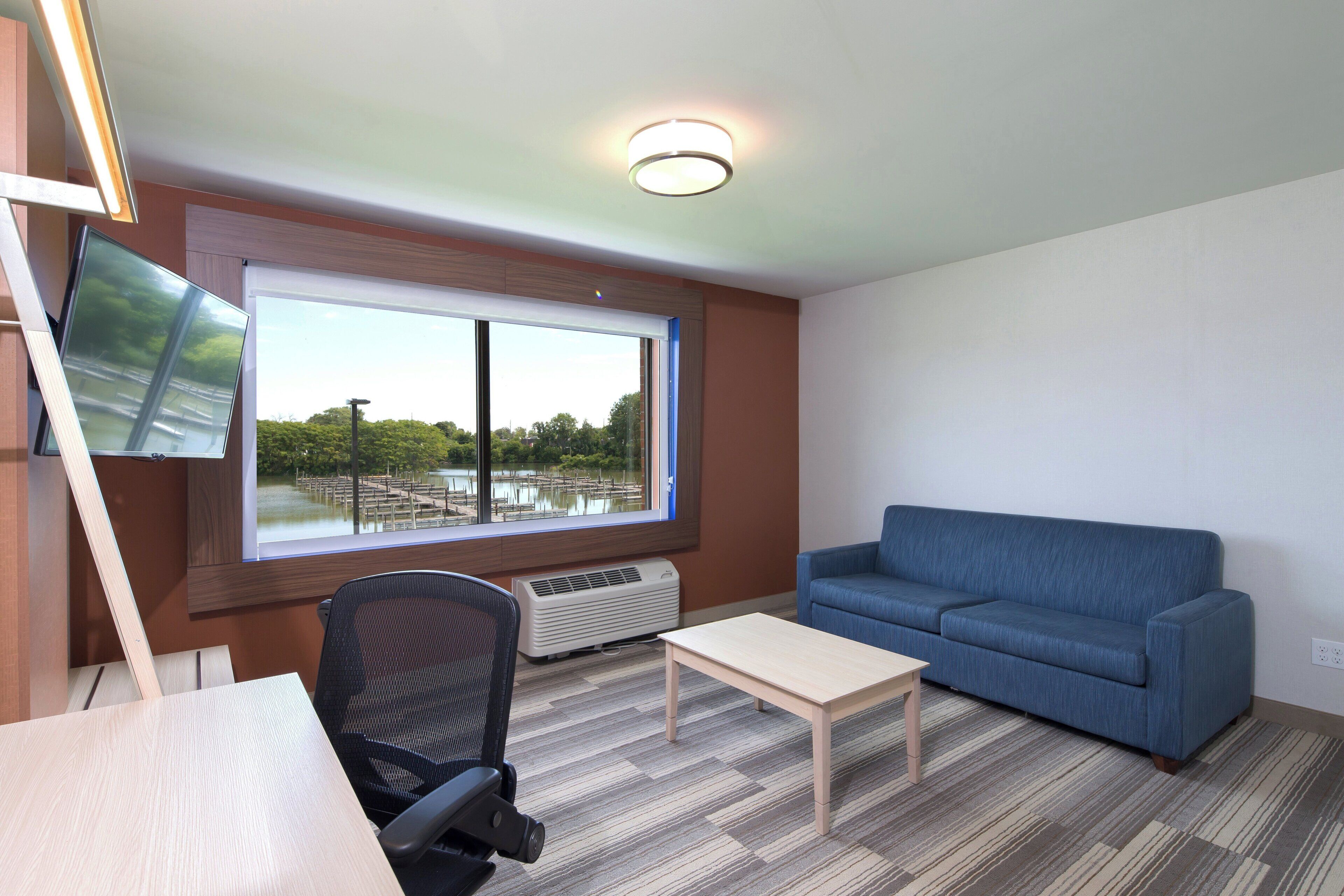 A room with a large window, a blue sofa, a small table, a chair, and a flat-screen TV mounted on the wall.