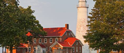 A lighthouse with a brick building in front, surrounded by trees.