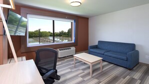 A room with a large window, a blue sofa, a wooden table, a black office chair, and a ceiling light fixture. - Holiday Inn Express & Suites Port Huron by IHG (Port Huron)
