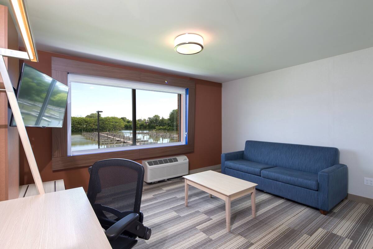 Ein Zimmer mit großem Fenster, einem blauen Sofa, einem Holztisch, einem schwarzen Bürostuhl und einer Deckenleuchte.
