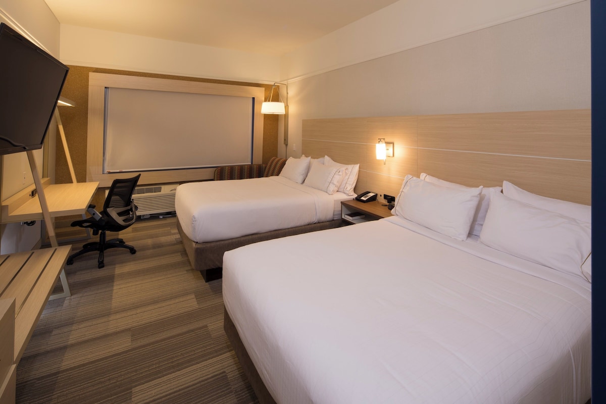 Une chambre d’hôtel avec deux lits, un bureau, une chaise, une télévision et une lampe fixée au mur.