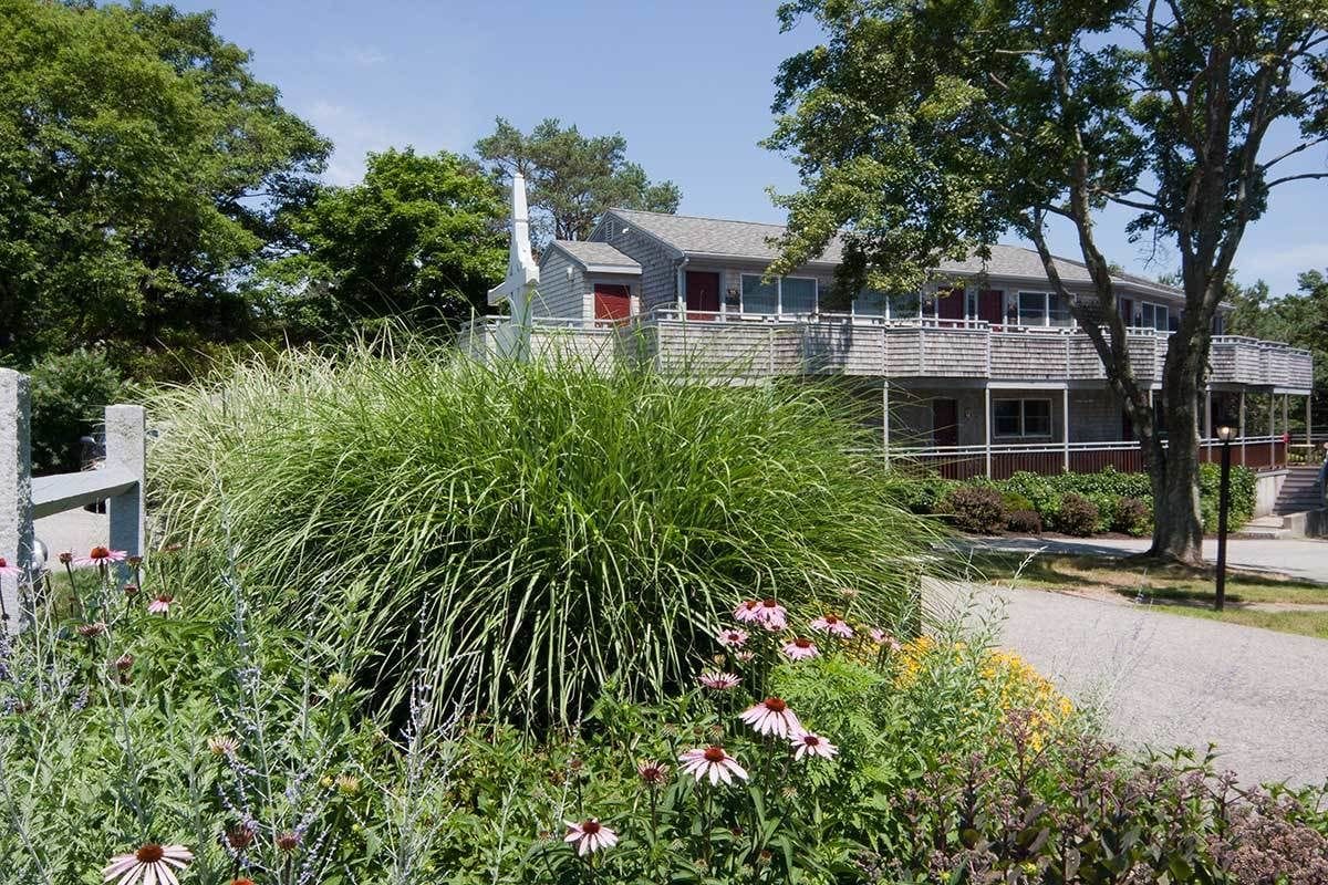 Un bâtiment avec une porte rouge, entouré d’un jardin où poussent différentes plantes et fleurs.