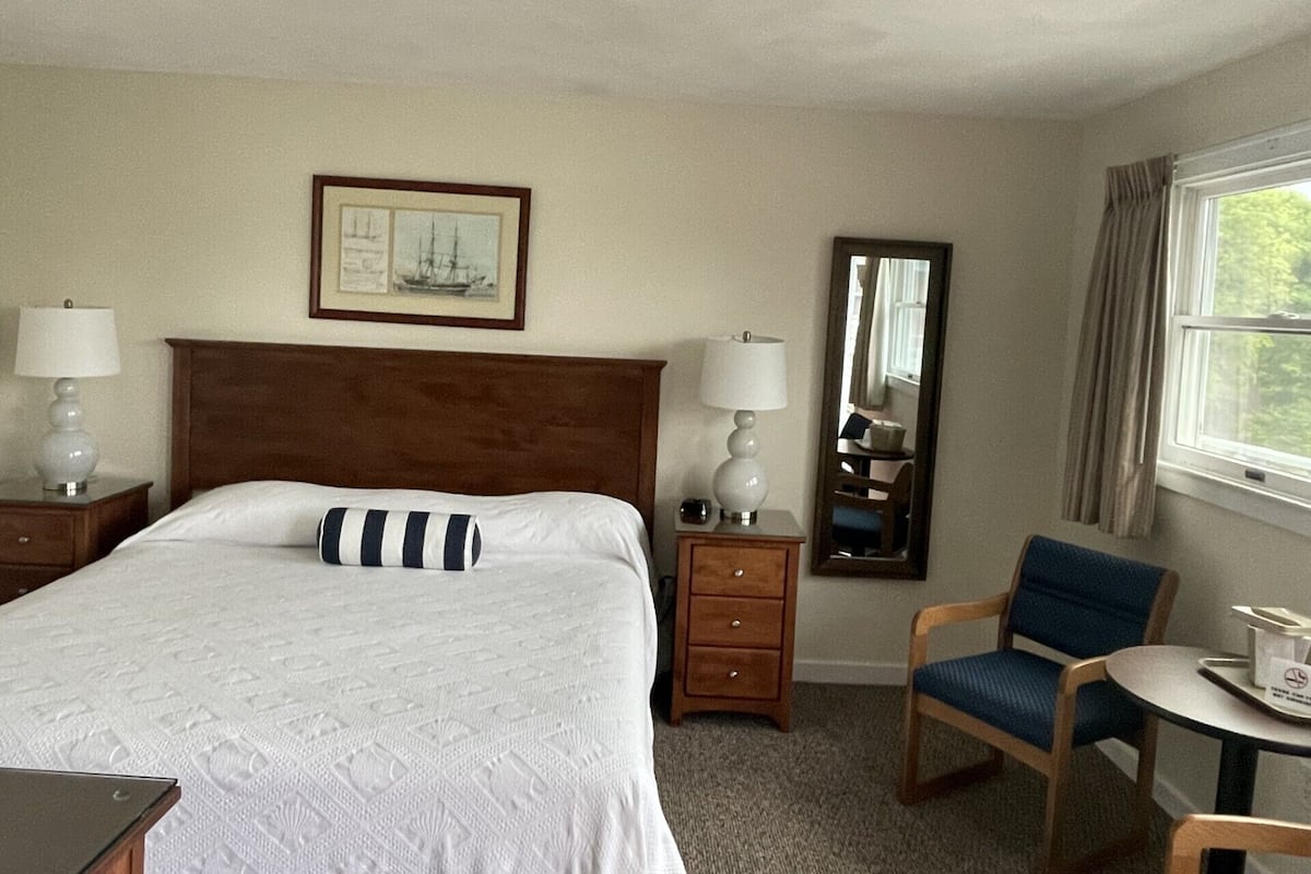 A hotel room with a large bed, two bedside tables with lamps, a small desk, a chair, a mirror, and a window with curtains.