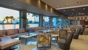 Un moderno vestíbulo de hotel con zona de sofás, mesas, sillas y grandes ventanales que ofrecen vistas a palmeras y montañas.