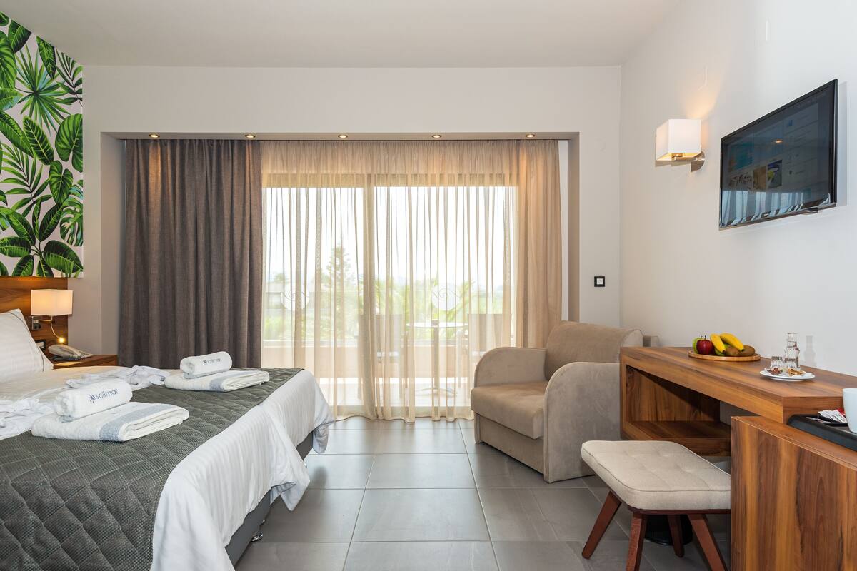 A hotel room with a bed, a desk, a chair, a bench, a potted plant, and a view of the outdoors.