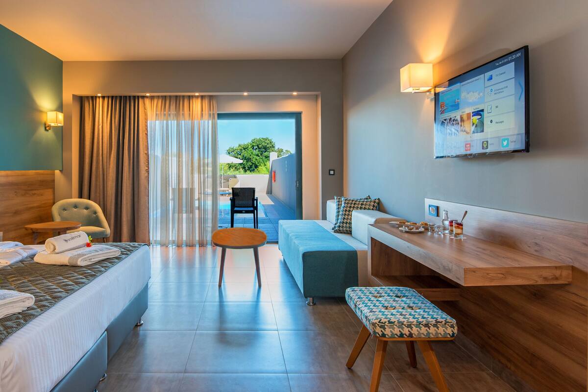 A hotel room with a bed, a desk, a chair, a small table, and a TV displaying a social media feed.